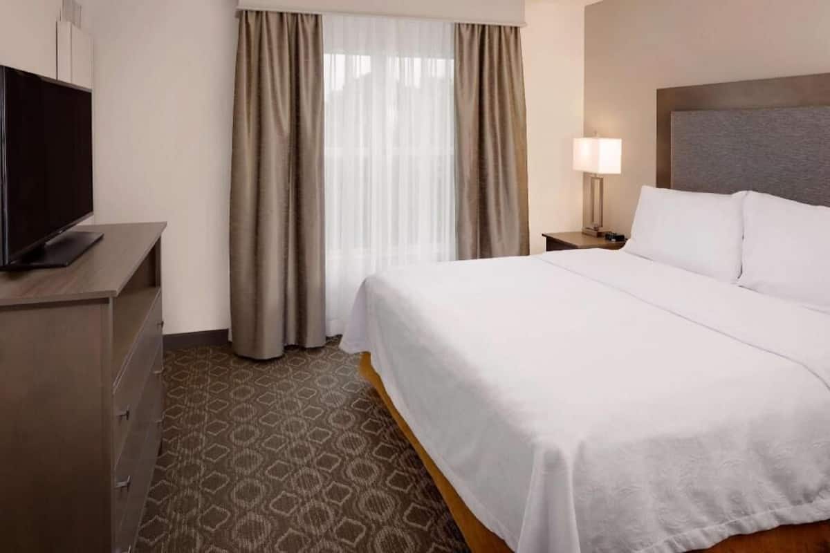 A hotel room with a bed, a TV on a dresser, a nightstand with a lamp, and a window with curtains.