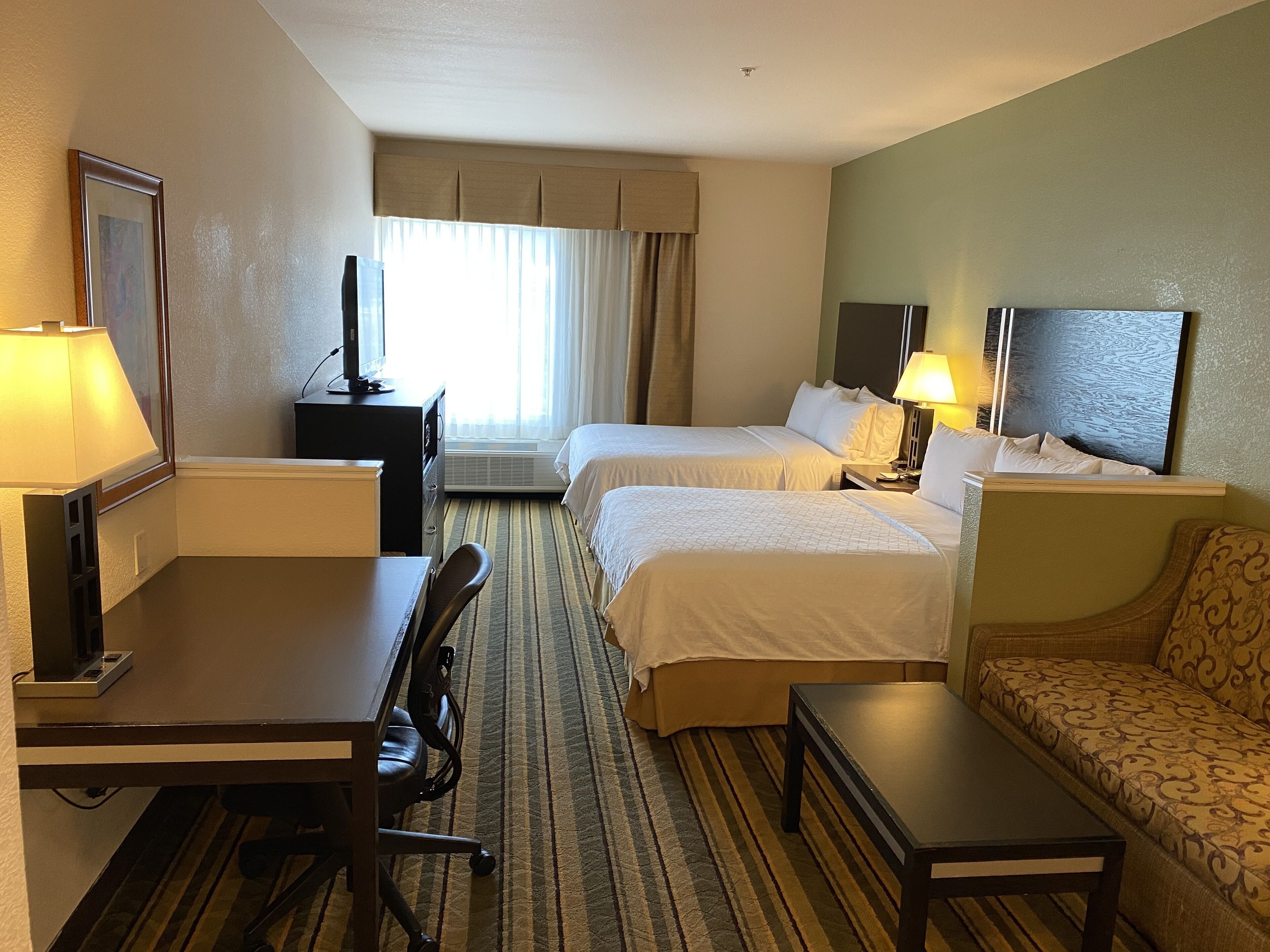A hotel room with two beds, a desk, a chair, a sofa, a TV, and a large window with curtains.