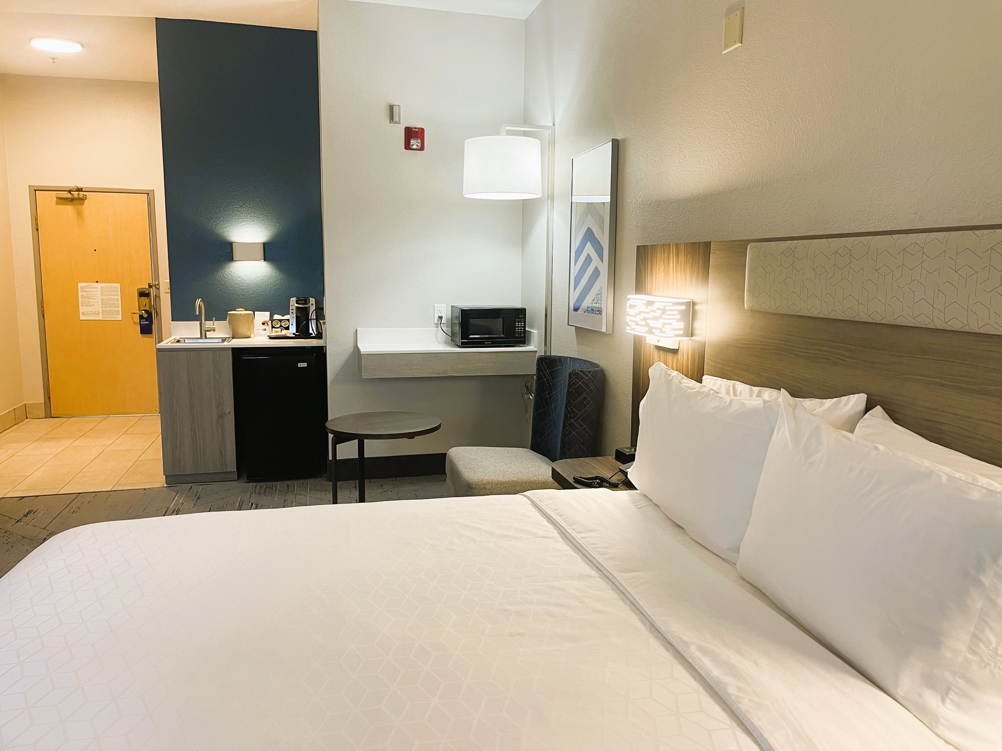 A hotel room with a bed, a desk with a microwave, a chair, and a small table.