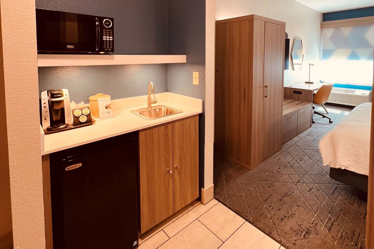 A compact hotel room with a kitchenette, a bed, and a view of the outdoors.