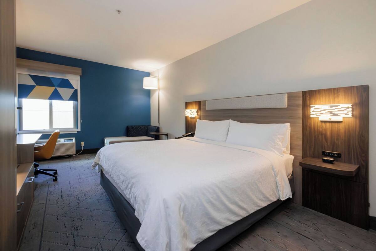 A hotel room with a large bed, a desk with a chair, a desk lamp, and a nightstand with a lamp.