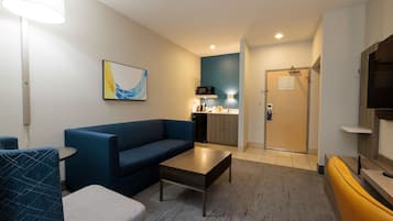 A modern hotel room with a blue sofa, a small table, a TV, and a kitchen area with a microwave.