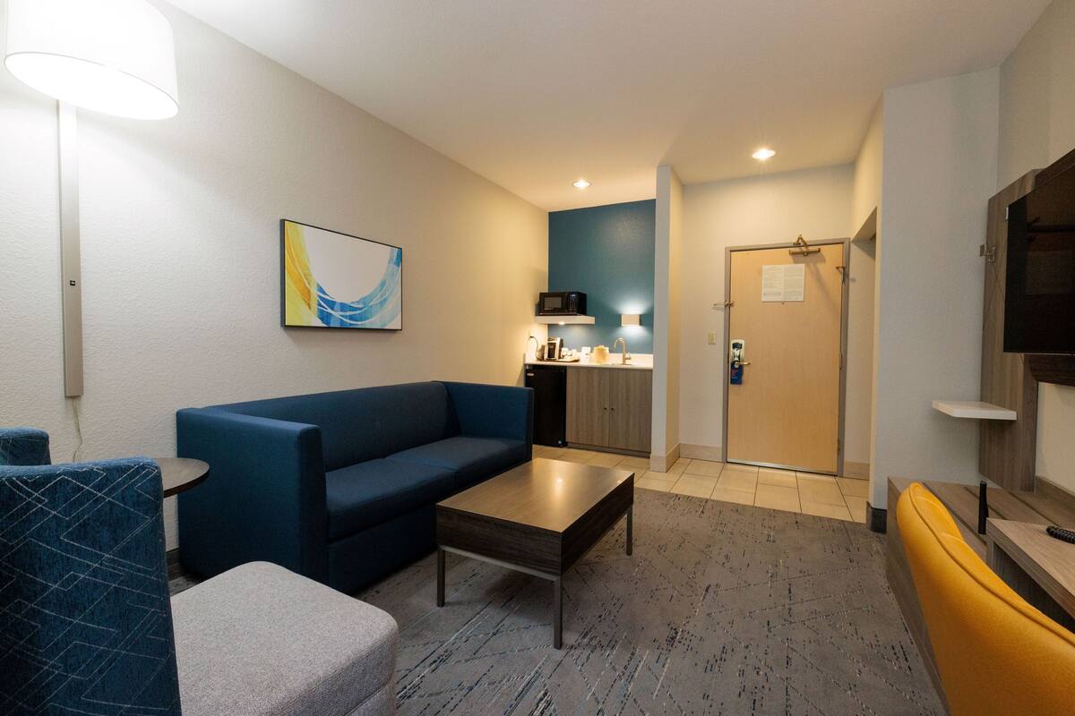 A modern hotel room with a blue sofa, a small table, a TV, and a kitchen area with a microwave.