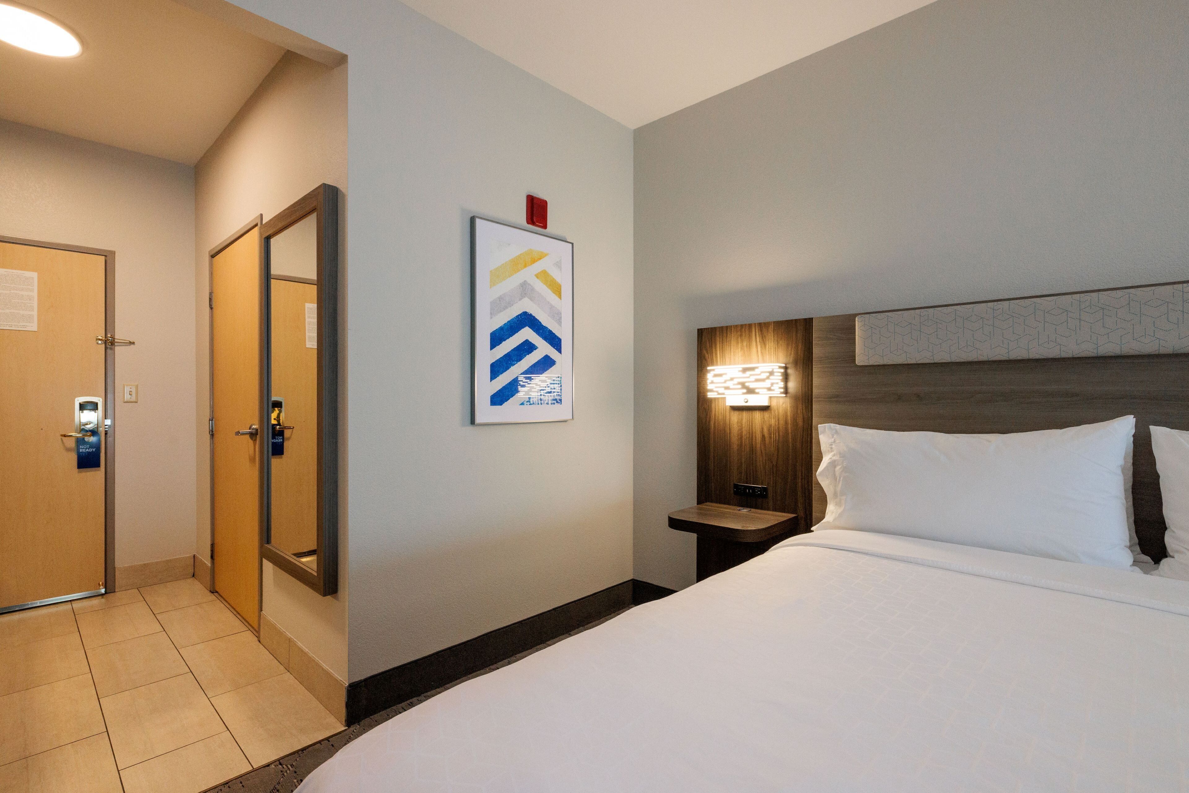 A hotel room with a bed, a nightstand, a wall-mounted light, a door, and a framed picture.