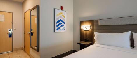 A hotel room with a bed, a nightstand, a wall-mounted light, a door, and a framed picture.