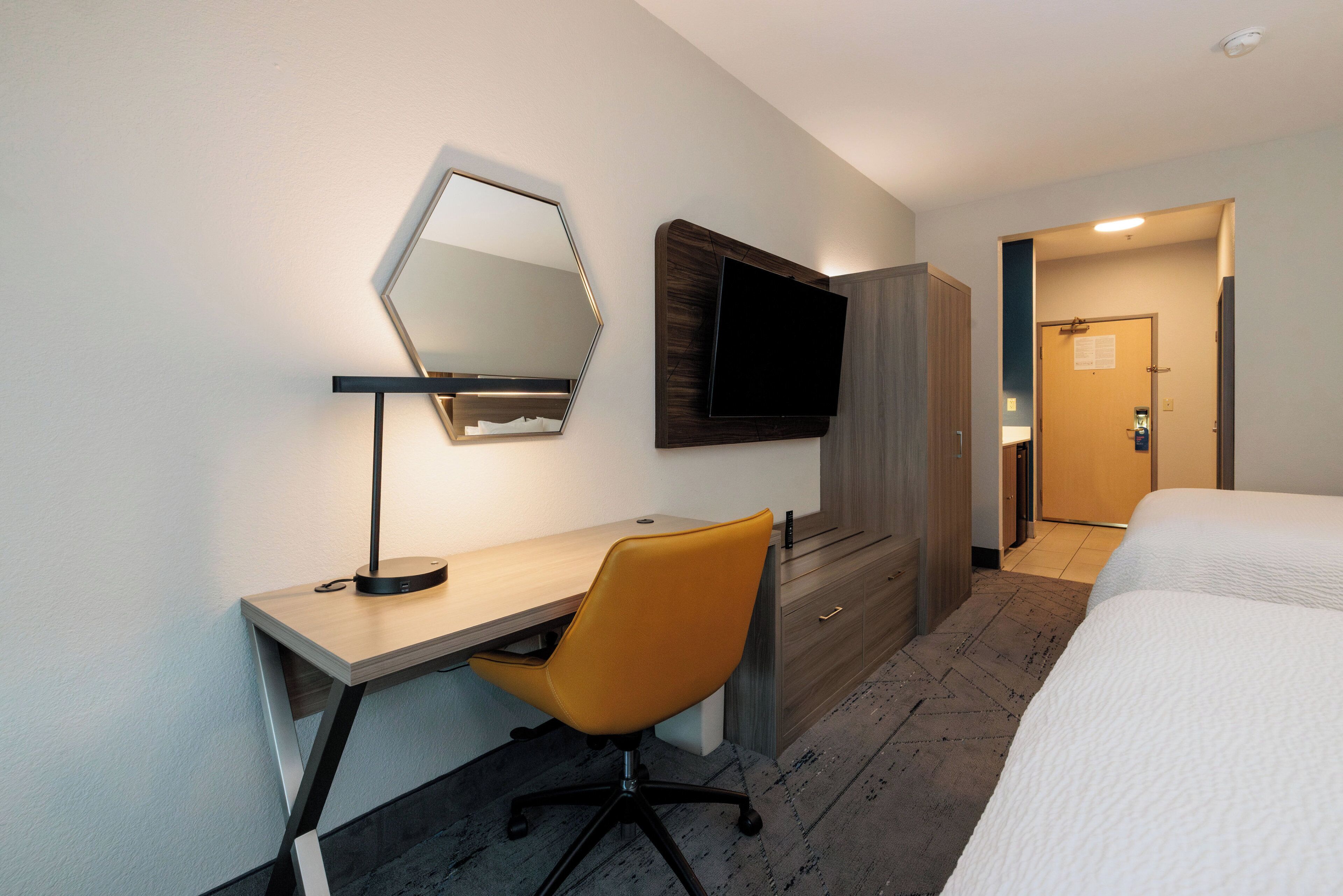 Habitación de hotel con escritorio de madera, una silla moderna, televisor de pantalla plana en la pared, cama con ropa de cama blanca y una puerta que da a otra habitación.
