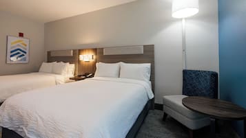 Quarto de hotel com duas camas, uma mesa de cabeceira com abajur, uma cadeira e uma mesinha.