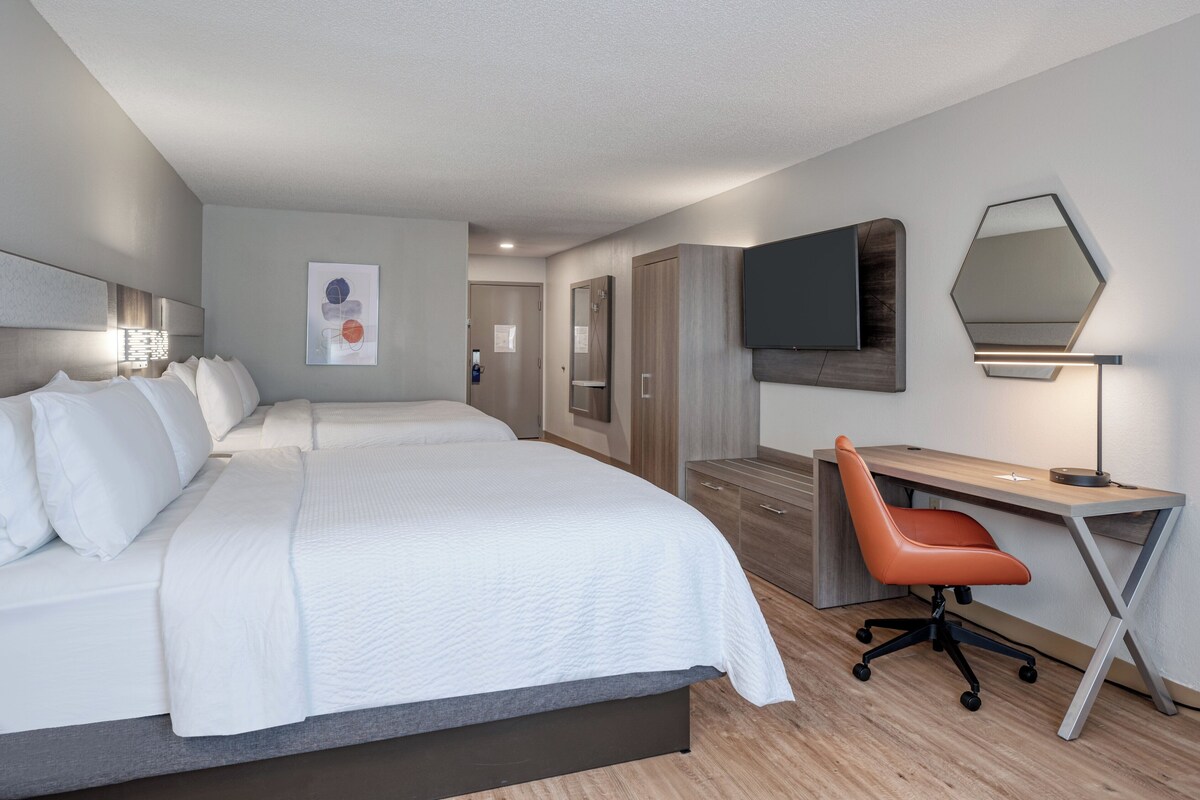 Quarto de hotel moderno com uma cama grande, uma escrivaninha e uma cadeira. Há uma TV de tela plana na parede e uma obra de arte emoldurada acima da cama.