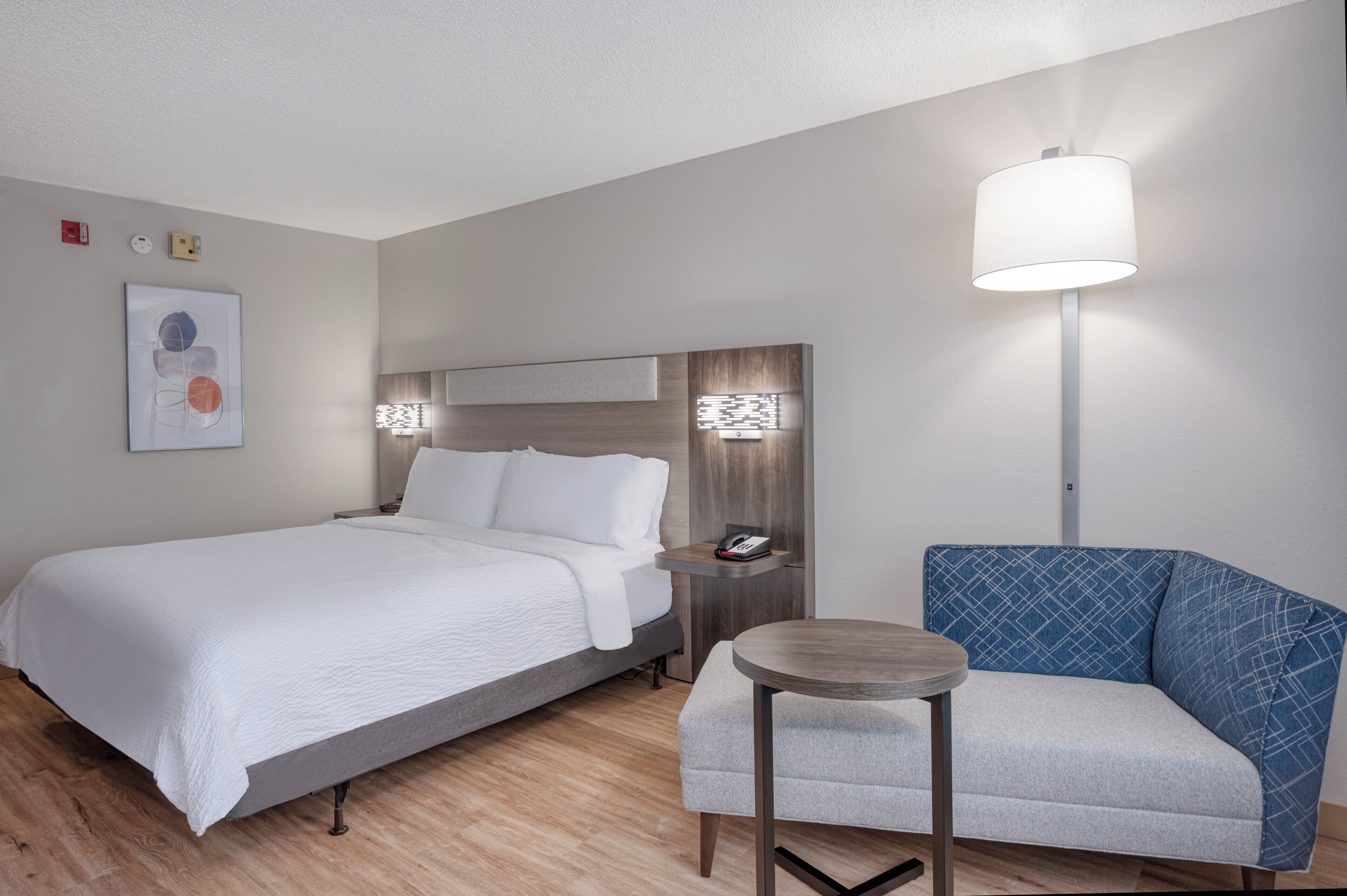 Una habitación de hotel con cama, mesita de noche, lámpara, silla y una obra de arte en la pared.