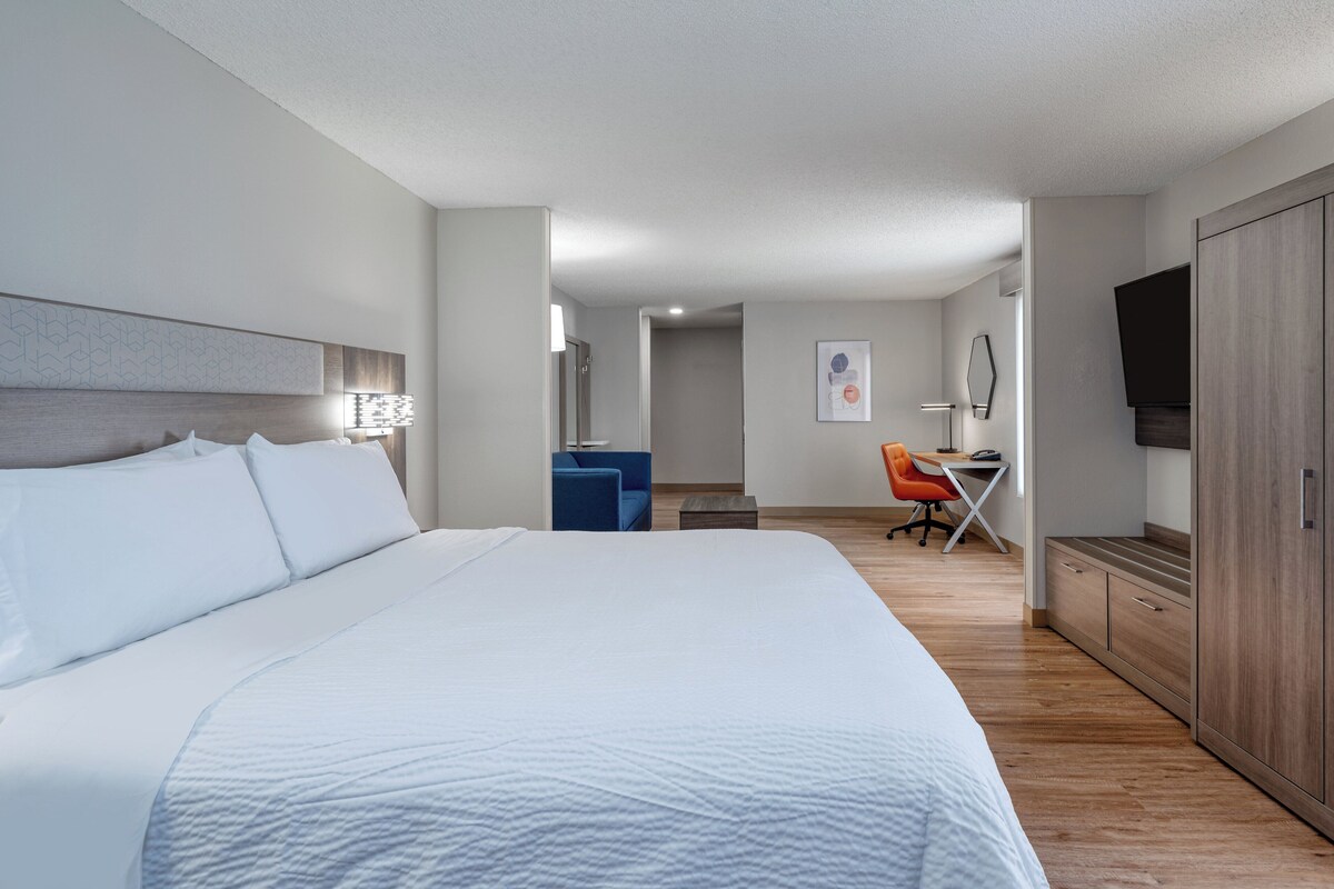 Quarto de hotel com uma cama grande, uma escrivaninha com cadeira, um guarda-roupa e uma televisão na parede.