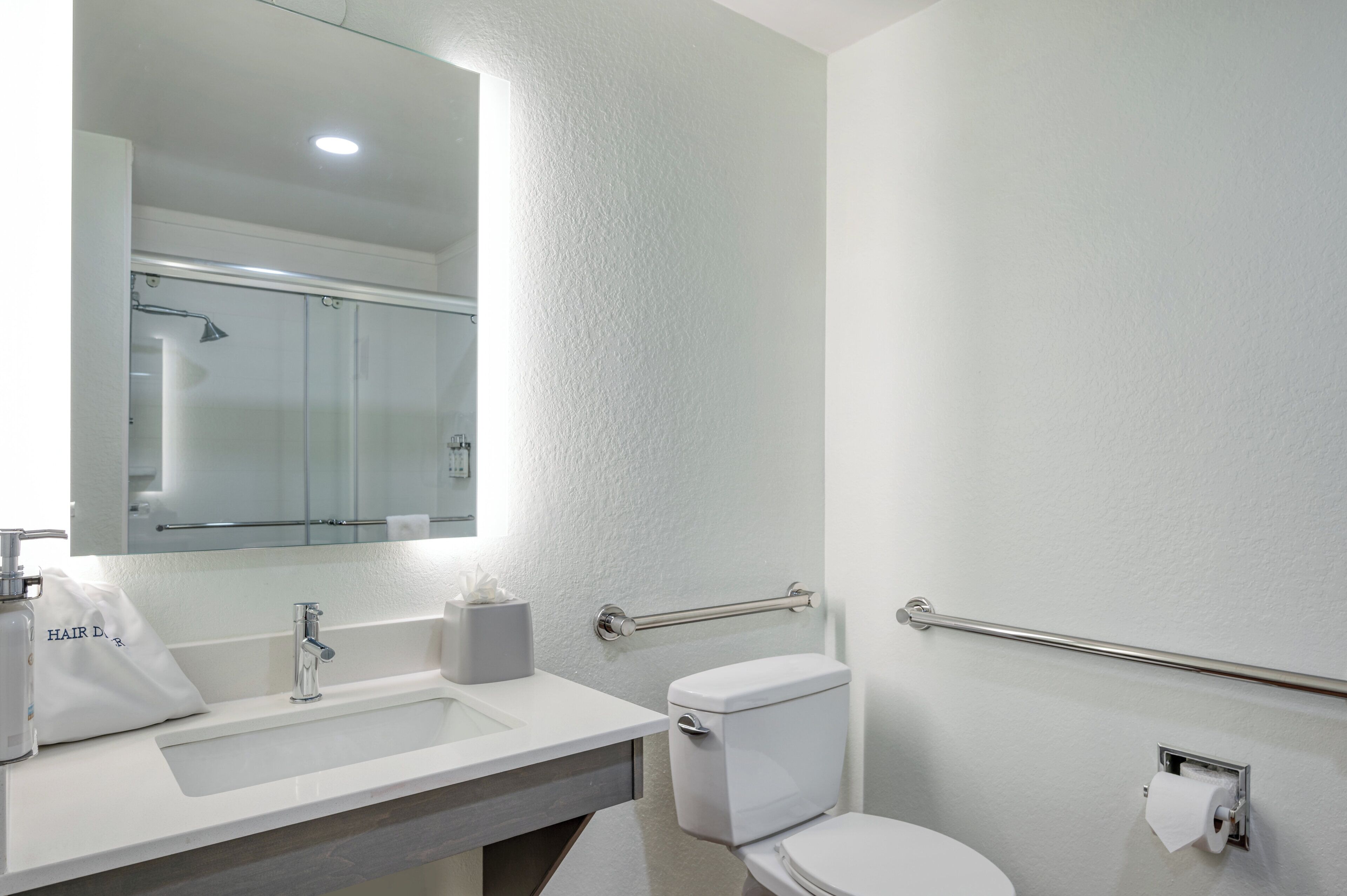 Baño con lavamanos blanco, espejo, inodoro y barras de apoyo.