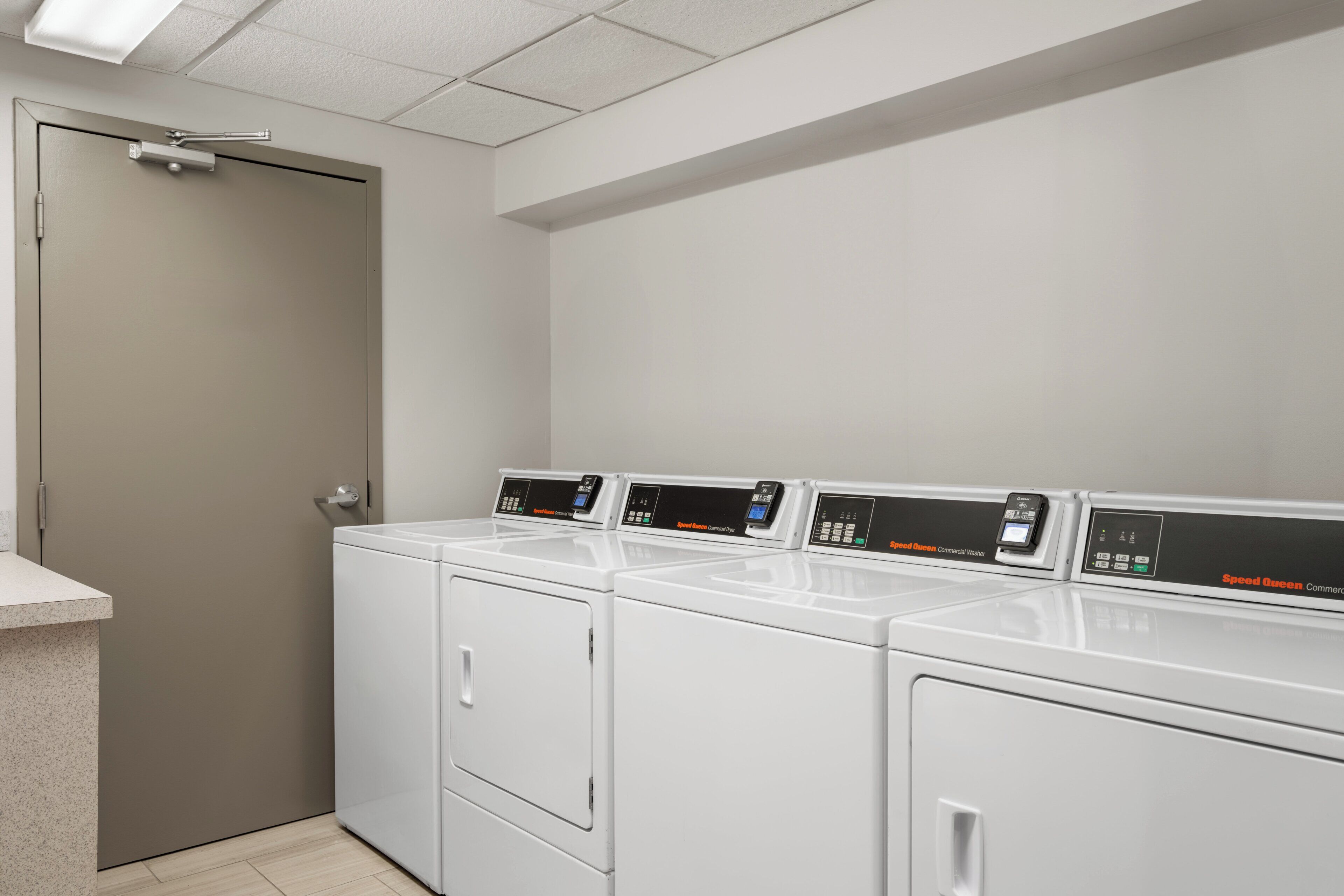 A laundry room with four white front-loading washers and dryers, a door, and a countertop.
