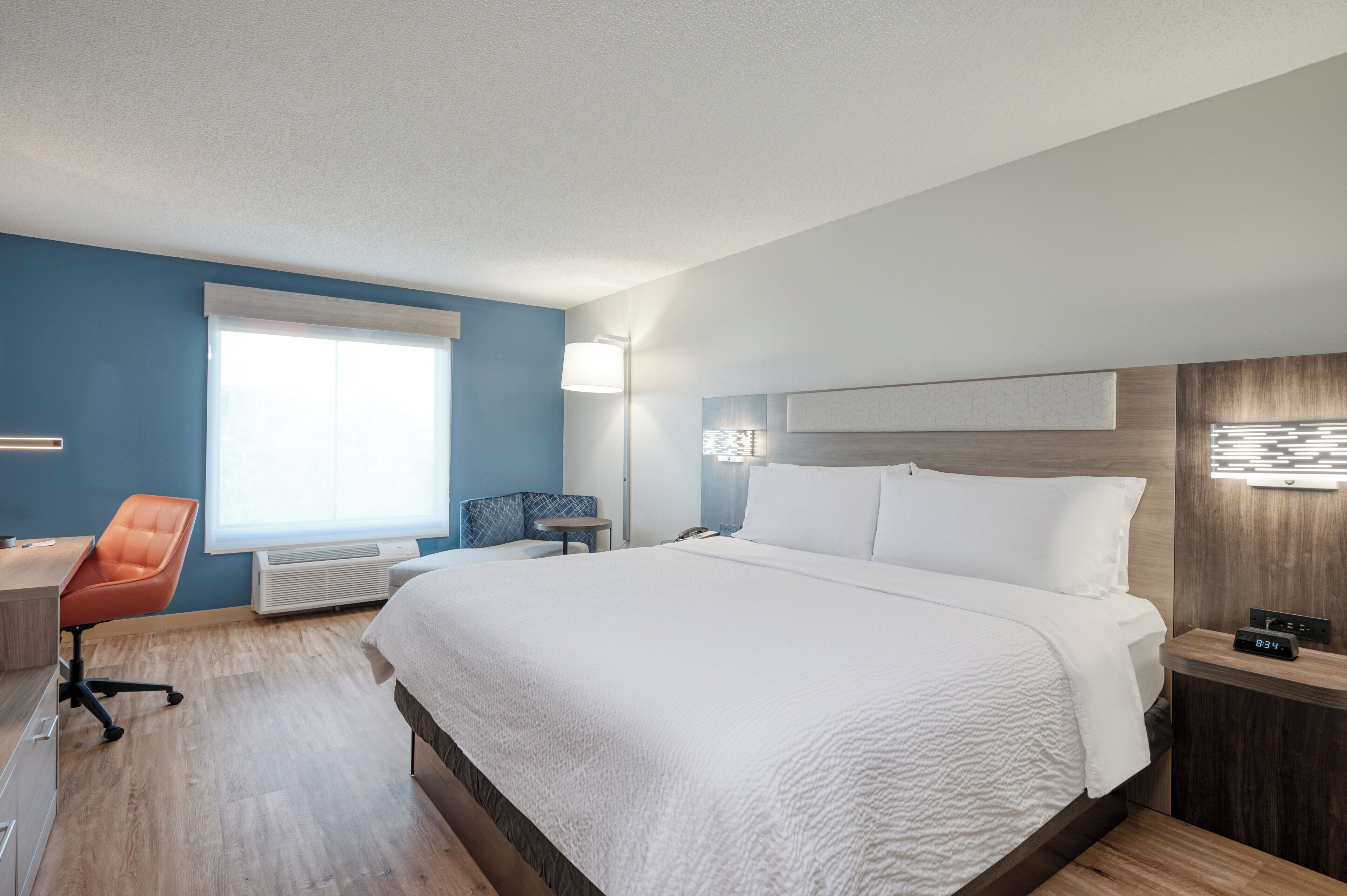 A hotel room with a large bed, a desk with a chair, a bedside table, and a window.