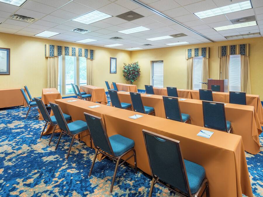 A conference room with rows of tables and chairs, large windows with curtains, and a carpeted floor.