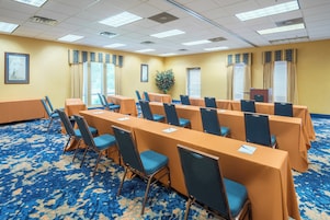 Un salón de conferencias con filas de mesas y sillas, ventanas amplias con cortinas y suelo alfombrado.