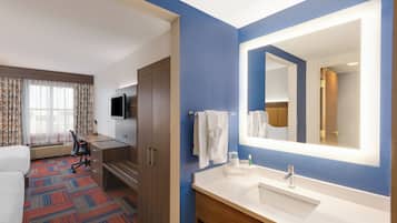 A hotel room with a bed, desk, chair, TV, bathroom with a sink and mirror, and a patterned carpet.