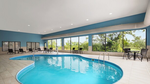 An indoor swimming pool with clear blue water, surrounded by black lounge chairs and tables, large windows offering a view of greenery outside. - Holiday Inn Express Morgantown by IHG (Morgantown)