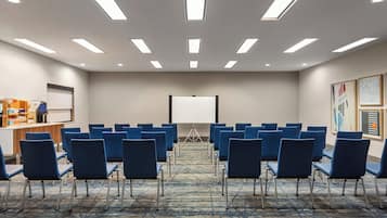 Un salón de conferencias con filas de sillas azules, una pantalla de proyector y un atril.