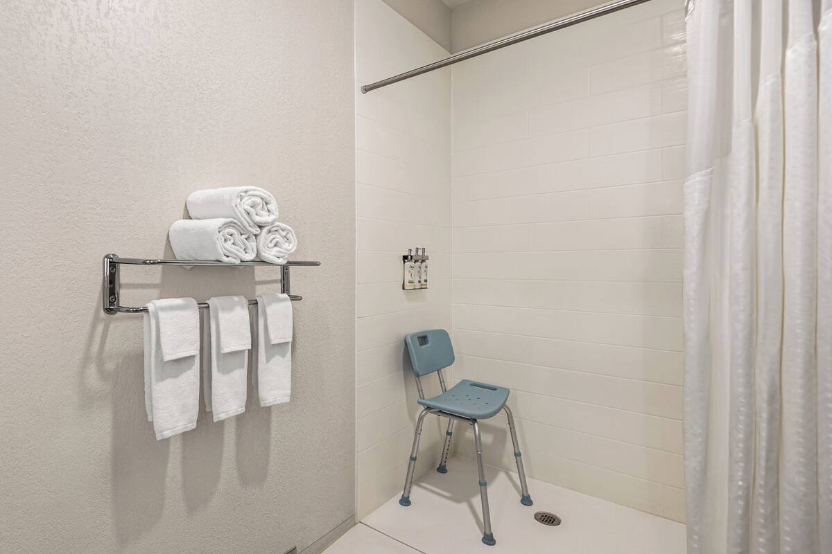 A bathroom with white tiled walls, a shower curtain, a towel rack with folded towels, and a wheelchair.