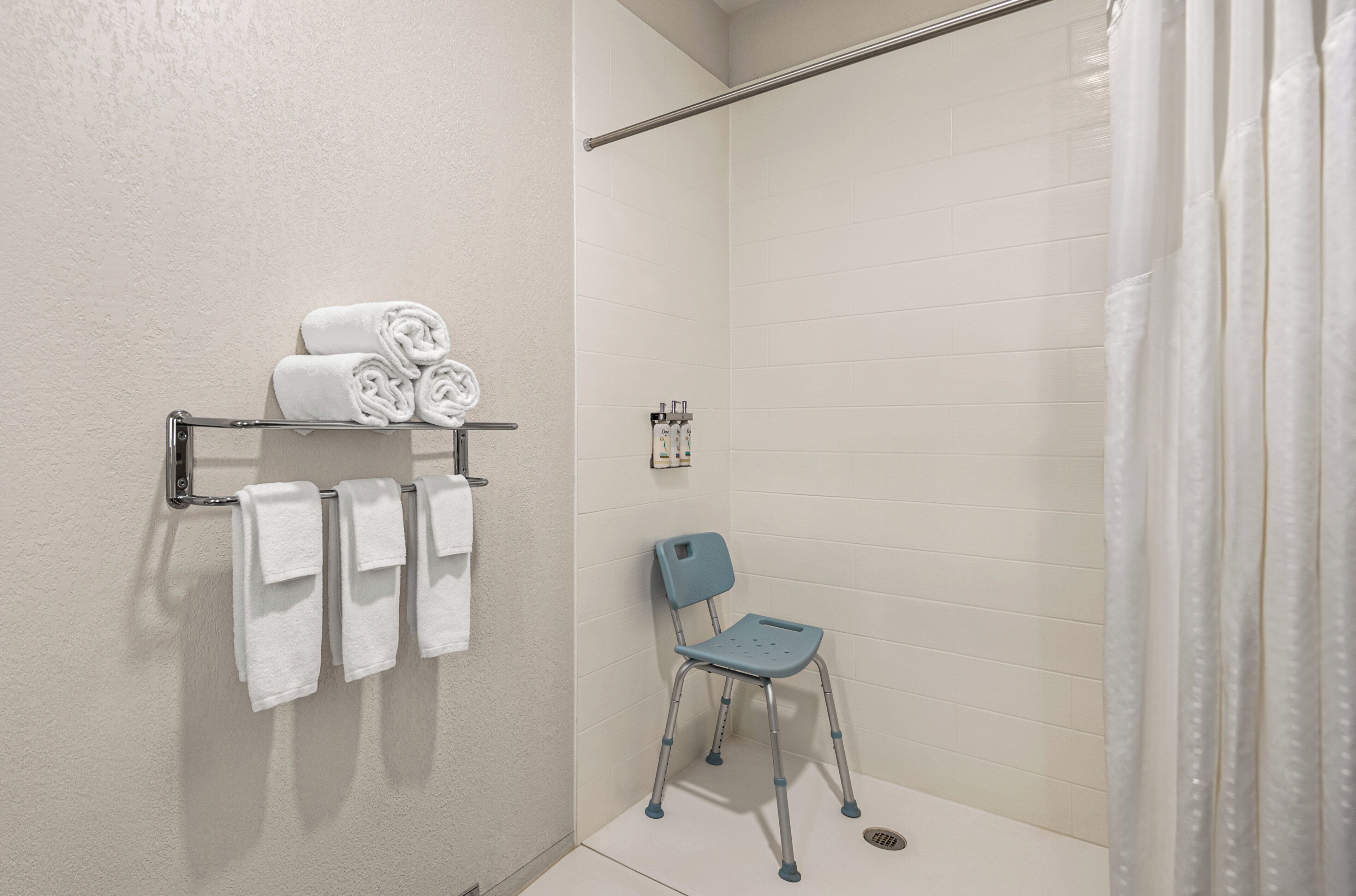 Un baño con paredes de azulejos blancos, cortina de ducha, toallero con toallas dobladas y una silla de ruedas.