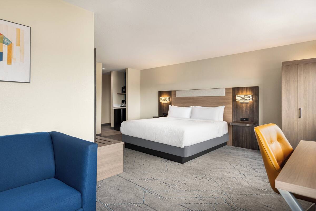 A modern hotel room with a large bed, a blue armchair, a wooden table, and a small desk with a lamp.