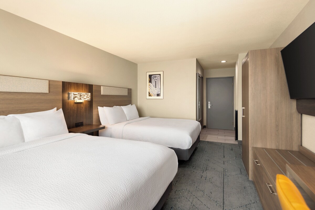 Ein Hotelzimmer mit zwei Betten, einem Flachbildfernseher, einem Holzschrank und einem gerahmten Kunstwerk an der Wand.