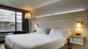 A hotel room with a large bed, a nightstand, a wall-mounted lamp, and a view of the outside through a window. - Holiday Inn Express Milwaukee North Brown Deer/Mequon by IHG (Milwaukee)