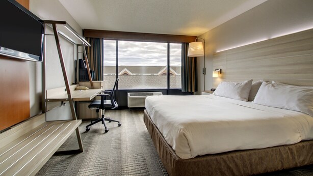 A hotel room with a large bed, a desk with a chair, a desk lamp, and a view of the cityscape through the window. - Holiday Inn Express Milwaukee North Brown Deer/Mequon by IHG (Milwaukee)