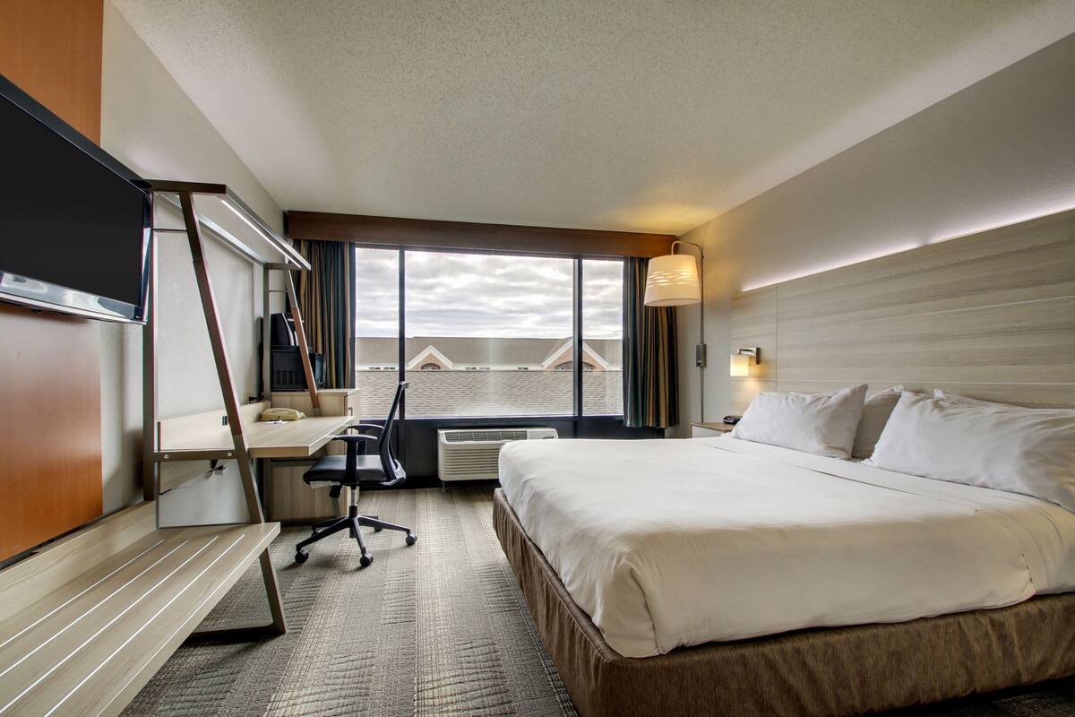 A hotel room with a large bed, a desk with a chair, a desk lamp, and a view of the cityscape through the window.