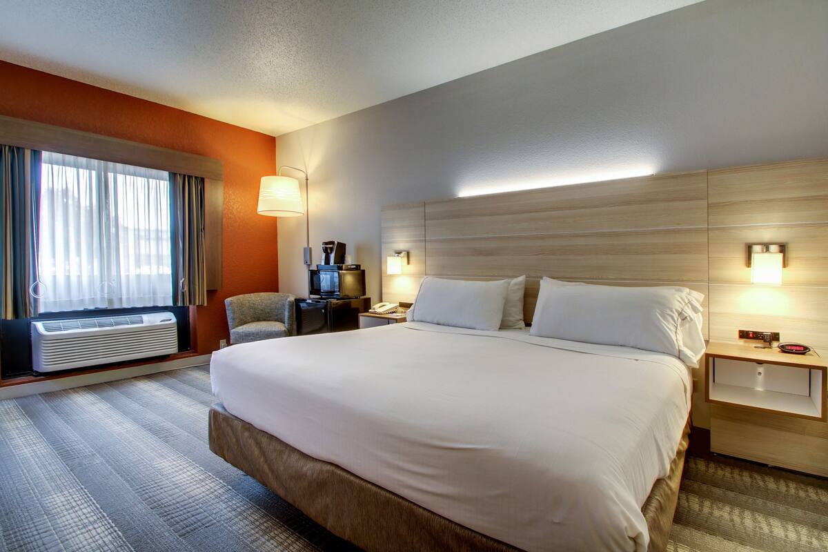A hotel room with a large bed, a desk with a lamp, a chair, a TV, and a window with curtains.
