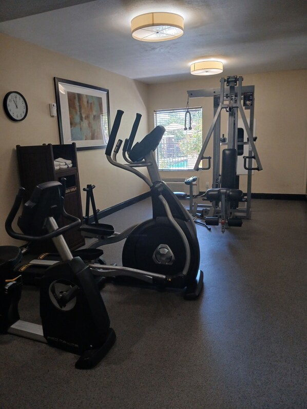 Une salle de sport équipée de machines de cardio, avec une horloge murale et une fenêtre munie de stores.