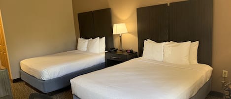 A hotel room with two single beds, a desk, a chair, a nightstand with a lamp, and a wall-mounted phone.