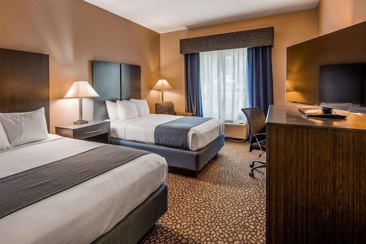 A hotel room with two beds, a desk, a chair, lamps, and a large window with curtains.