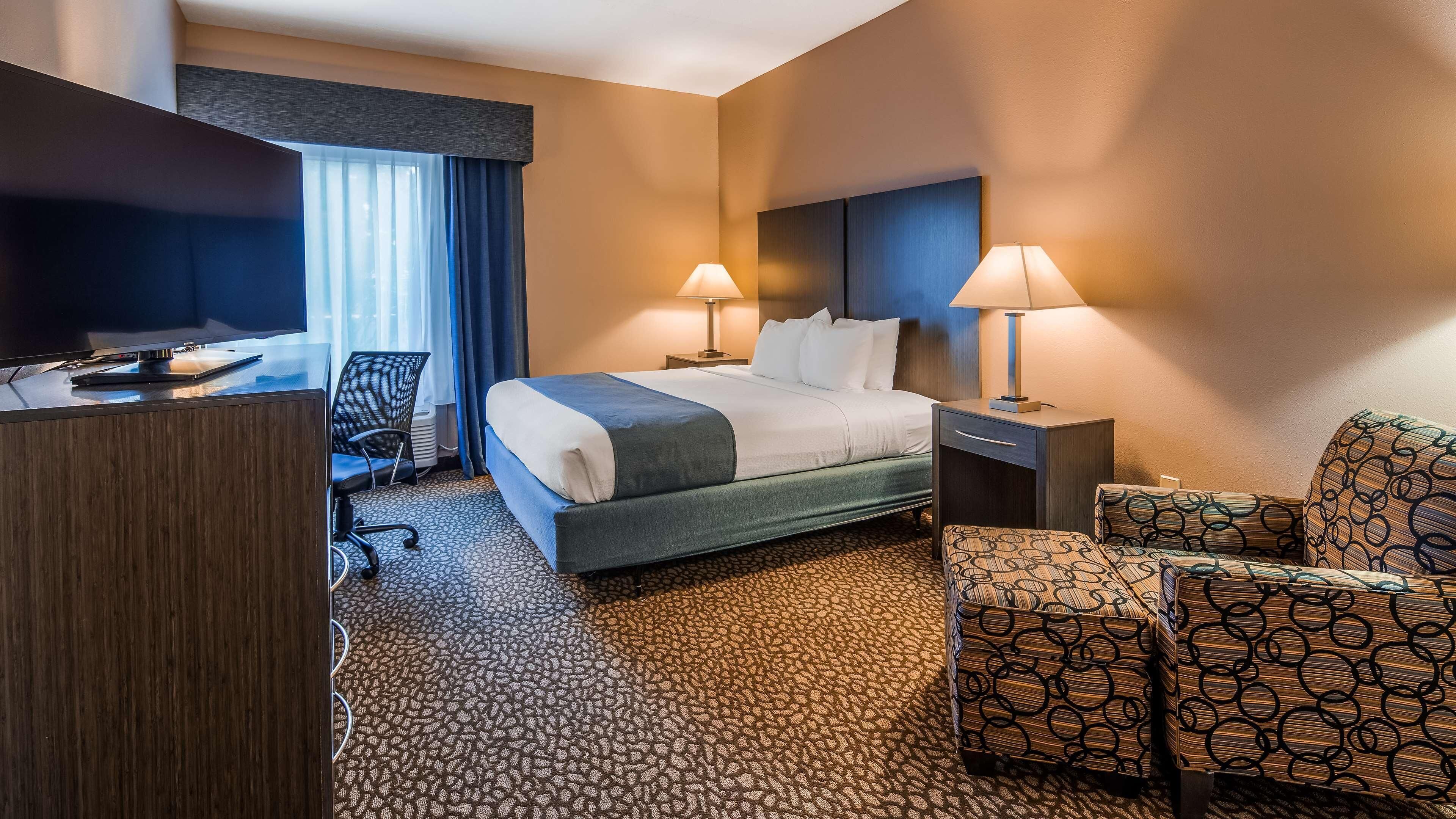 Une chambre d’hôtel comprenant un lit, deux lampes de chevet, un bureau avec une télévision, une chaise et un pouf à motifs.