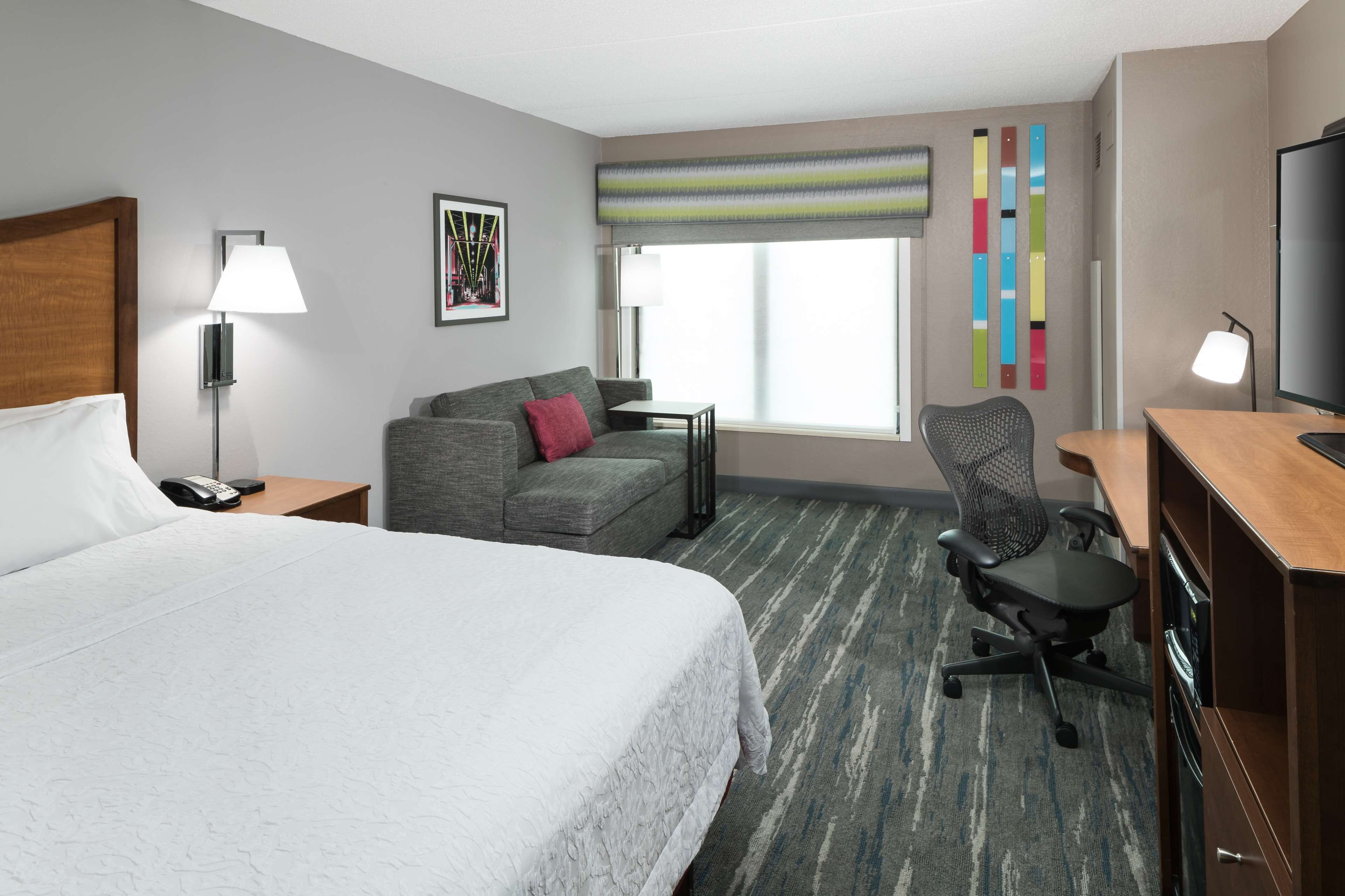 A hotel room with a large bed, a gray sofa, a desk with a chair, a TV, and a window with a view.