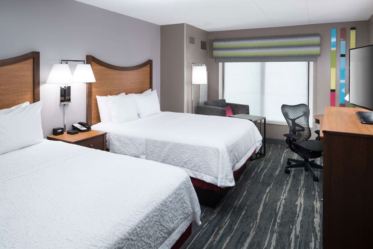 A hotel room with two beds, a desk, a chair, a lamp, and a window with a view.