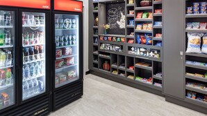 Una sección refrigerada con diversas bebidas y aperitivos, y una pared con estantes llenos de bocadillos y un letrero en una pizarra.