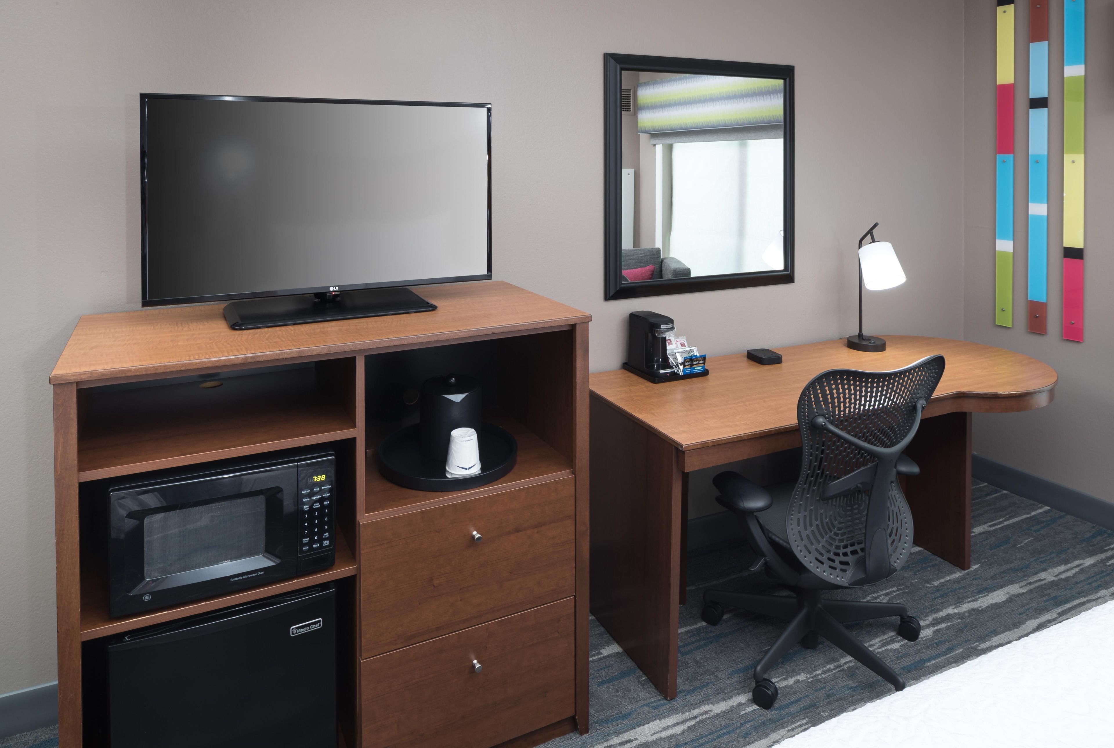 A hotel room with a flat-screen TV, a microwave, a coffee maker, a desk with a chair, and a mirror.