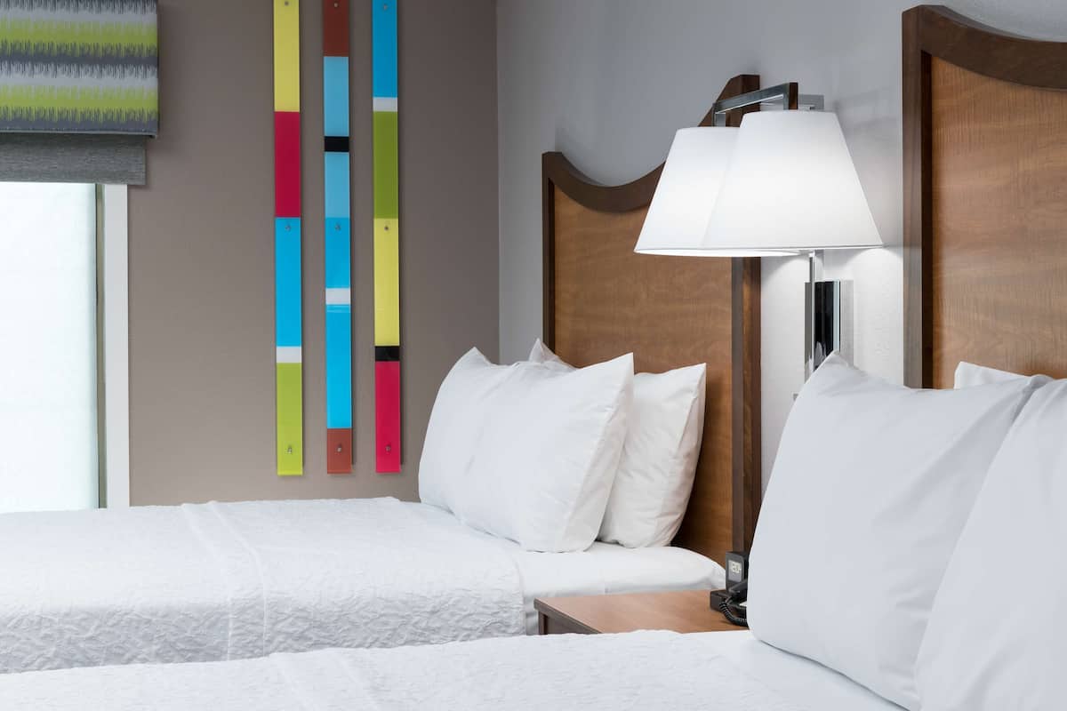 A hotel room with two beds, a bedside table, a lamp, and a colorful abstract painting on the wall.