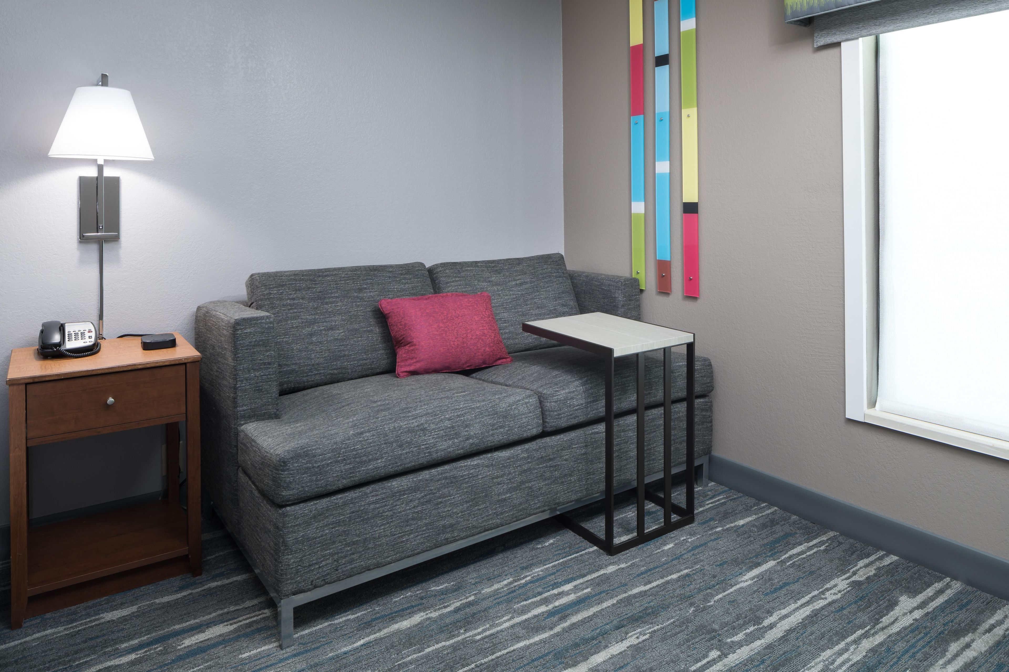 A modern hotel room with a grey sofa, a small table, a lamp, a phone, and a colorful abstract art piece on the wall.
