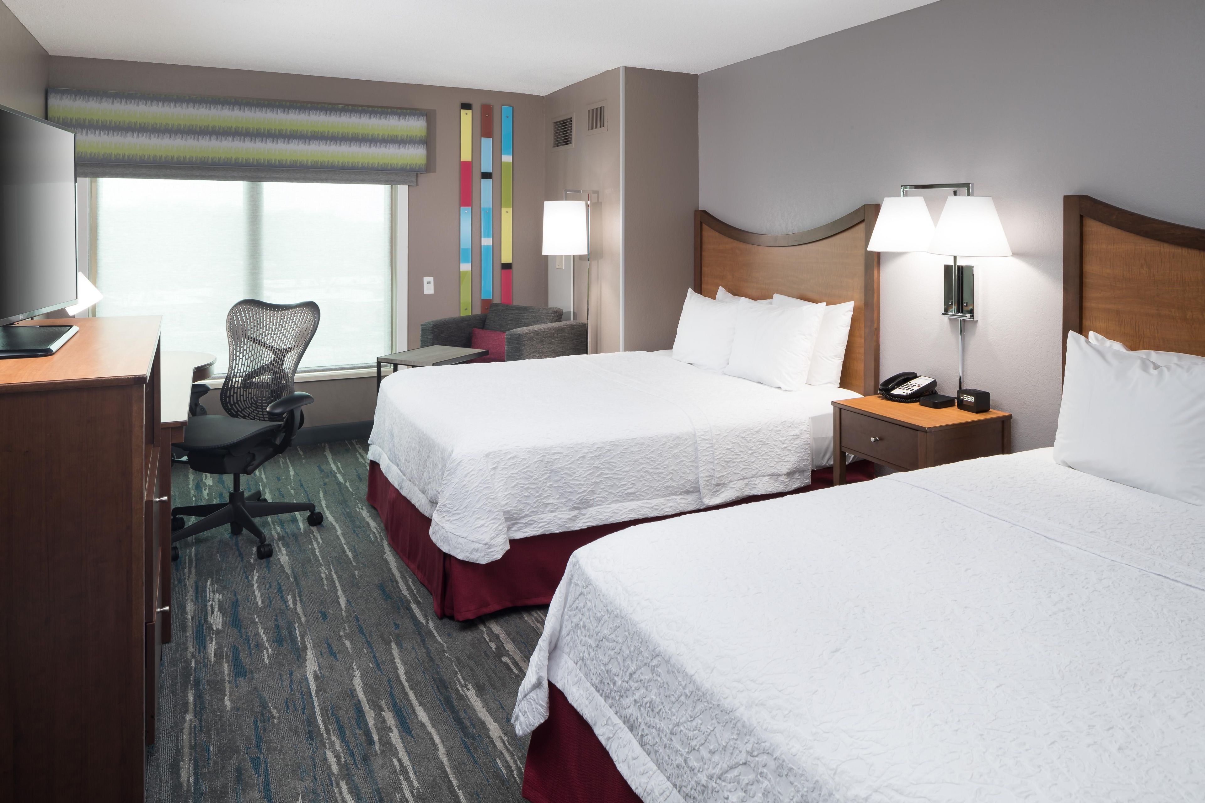 A hotel room with two beds, a desk, a chair, a lamp, and a phone.