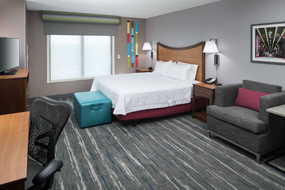A hotel room with a bed, a desk, a chair, a sofa, a lamp, a TV, and a window.