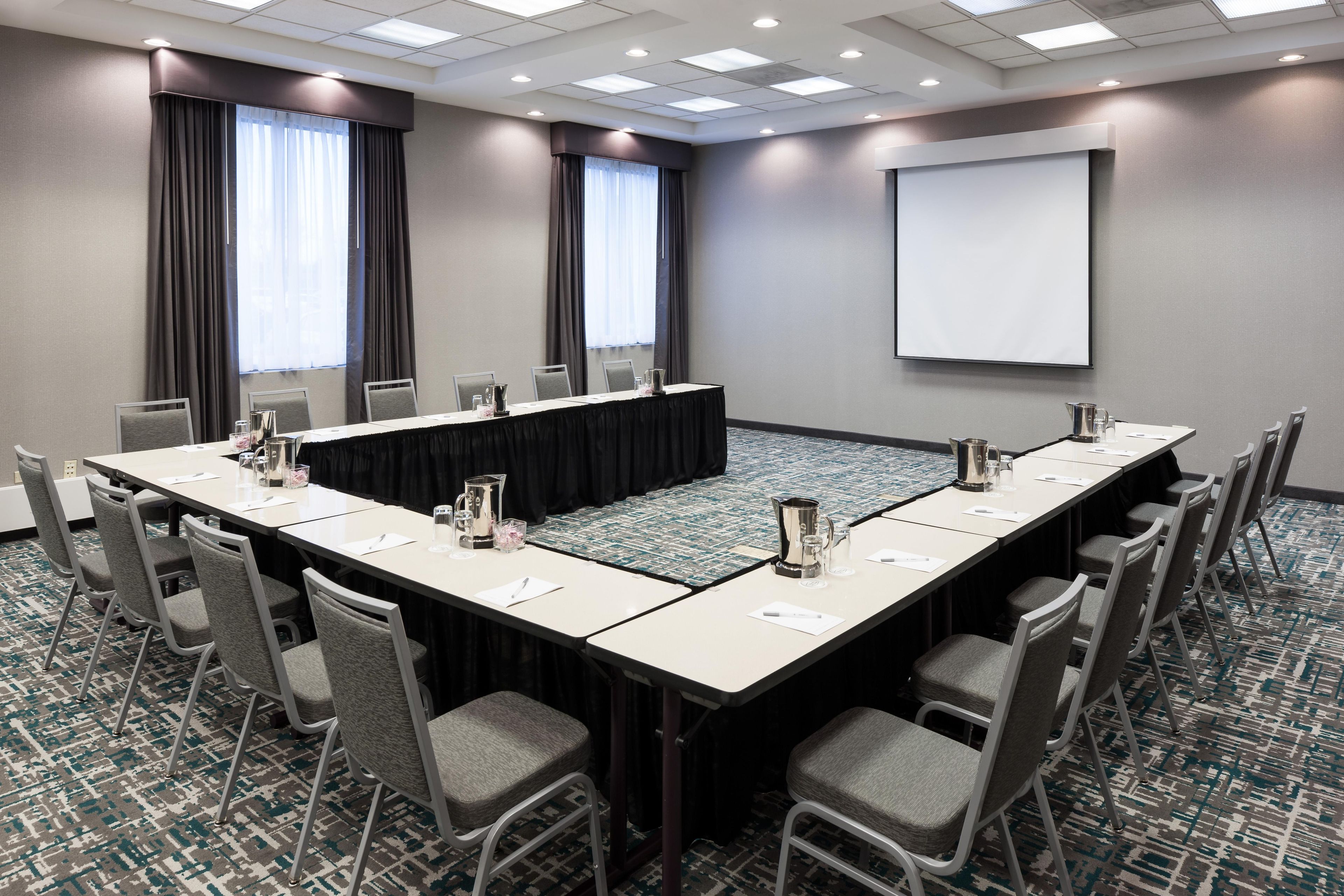 Un salón de conferencias con una larga mesa, sillas, una pantalla de proyector y piso alfombrado.