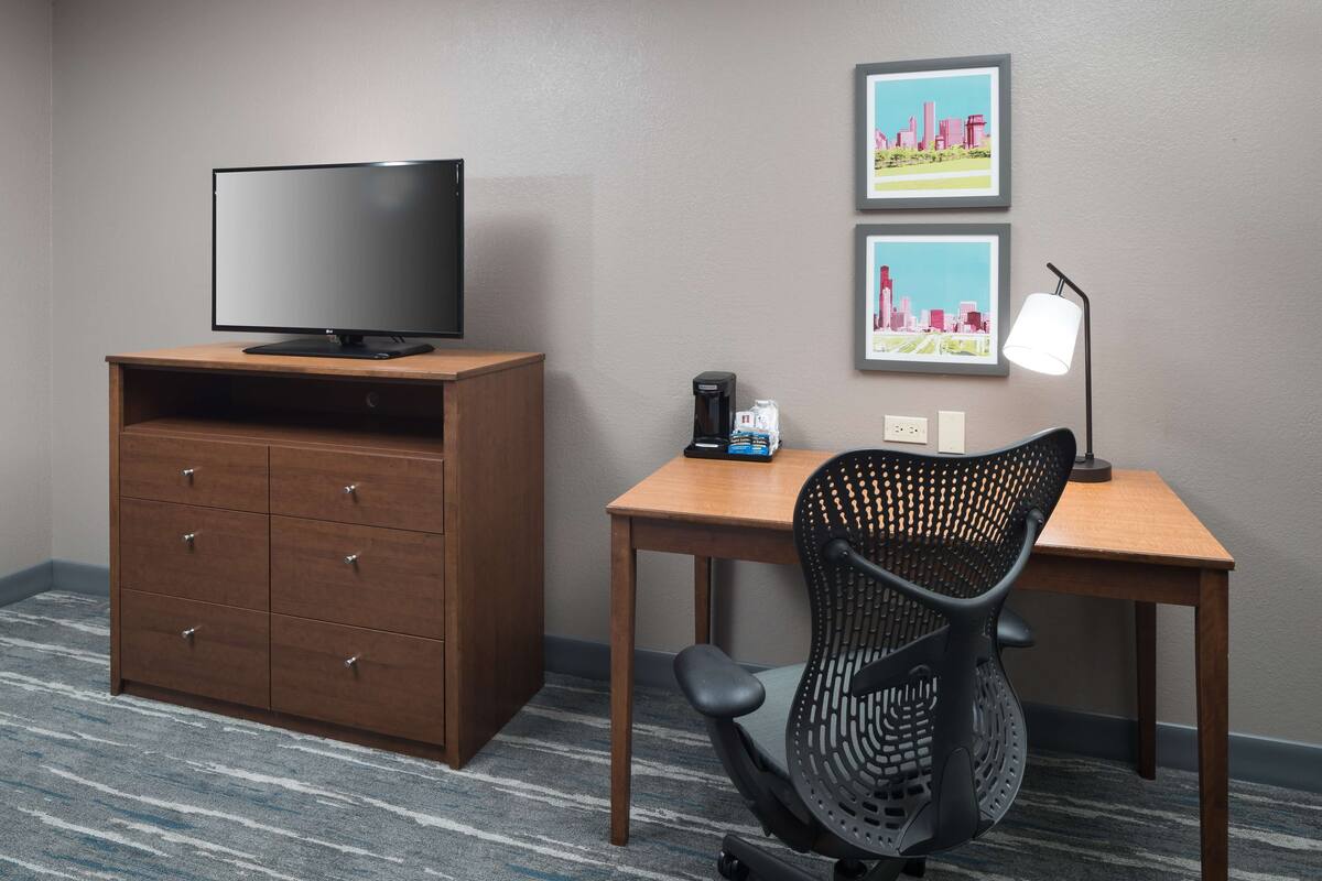 A hotel room with a wooden desk, a black office chair, a television on a wooden cabinet, and two framed pictures on the wall.