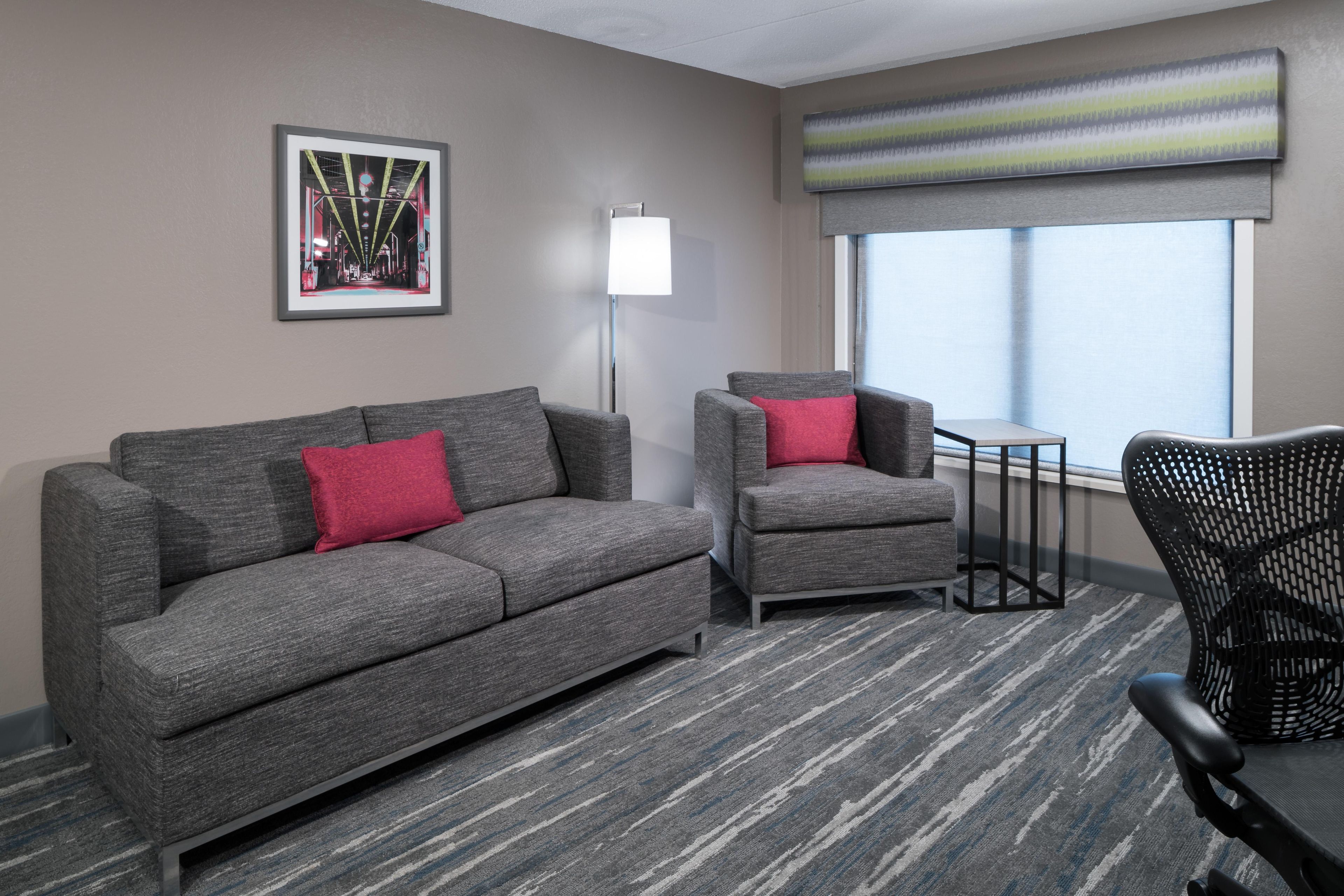 A hotel room with a gray sofa, a smaller armchair, a black chair, a small table, and a window with a view.