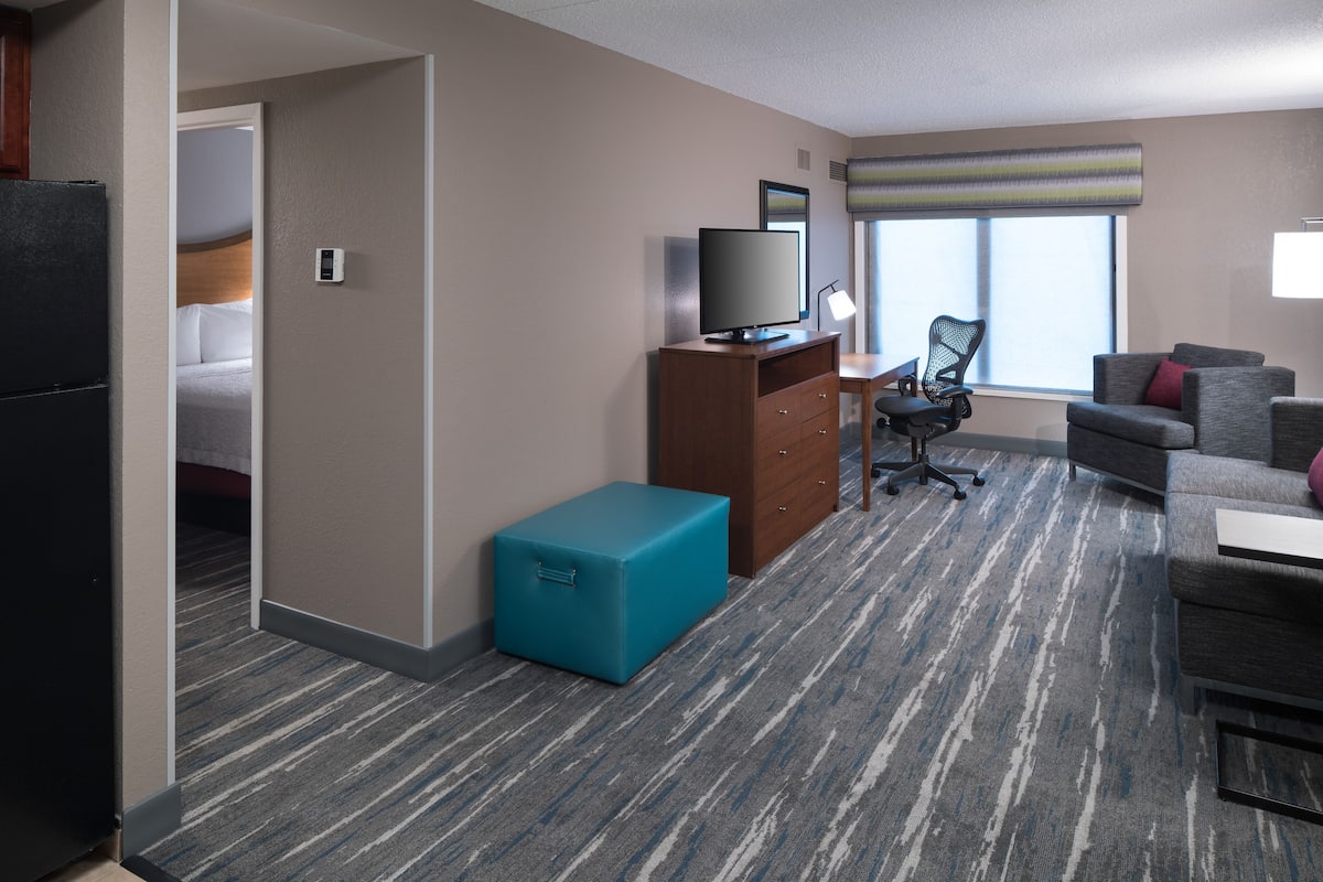 A hotel room with a bed, a desk with a computer, a TV, a chair, a sofa, and a small blue suitcase.