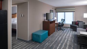 A hotel room with a bed, a desk with a computer, a TV, a chair, a sofa, and a small blue suitcase.