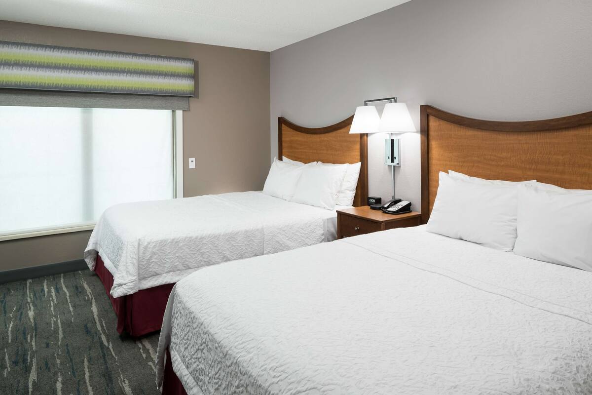 A hotel room with two beds, a nightstand with a lamp, and a window with blinds.