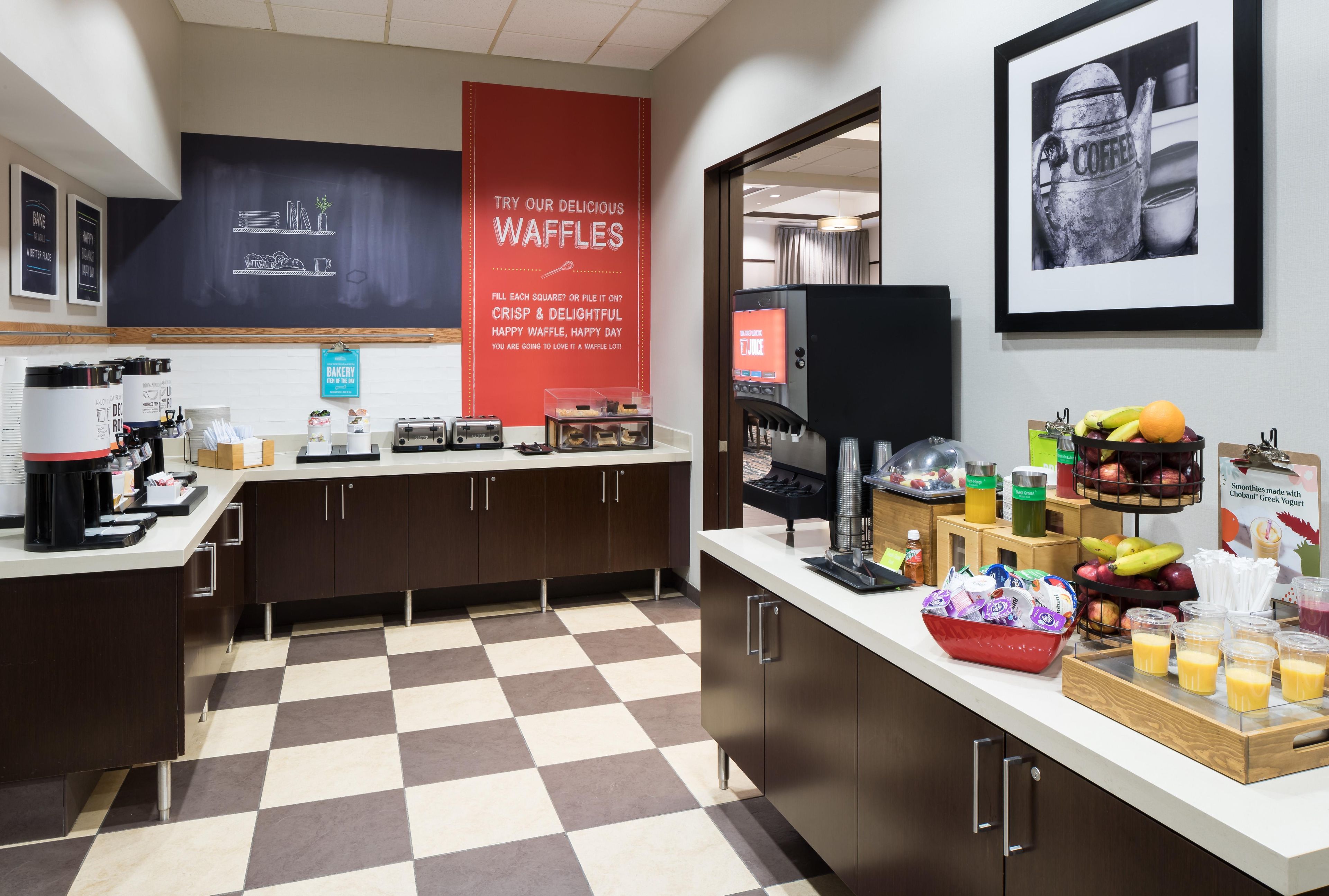 A modern kitchen area with a waffle station, coffee machines, and a juice dispenser.