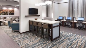 Un vestíbulo moderno de hotel con zona de bar, asientos y varias estaciones de trabajo con computadoras.